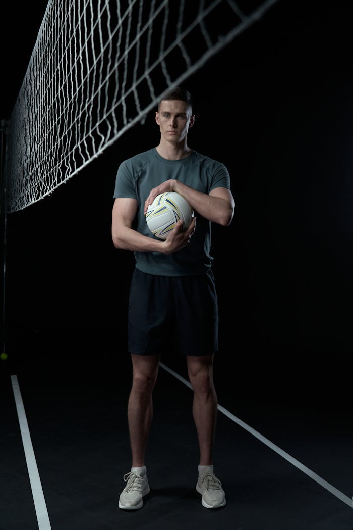 Athletic man in sportswear standing with a volleyball against a black background.