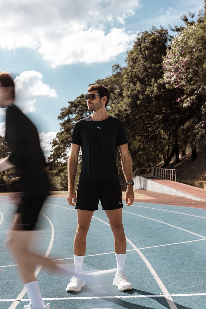 About Man in sports attire standing on an outdoor running track with blurred runner passing by.