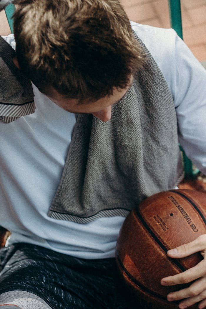 A basketball player taking a break, sitting with a towel over shoulders, holding a ball.