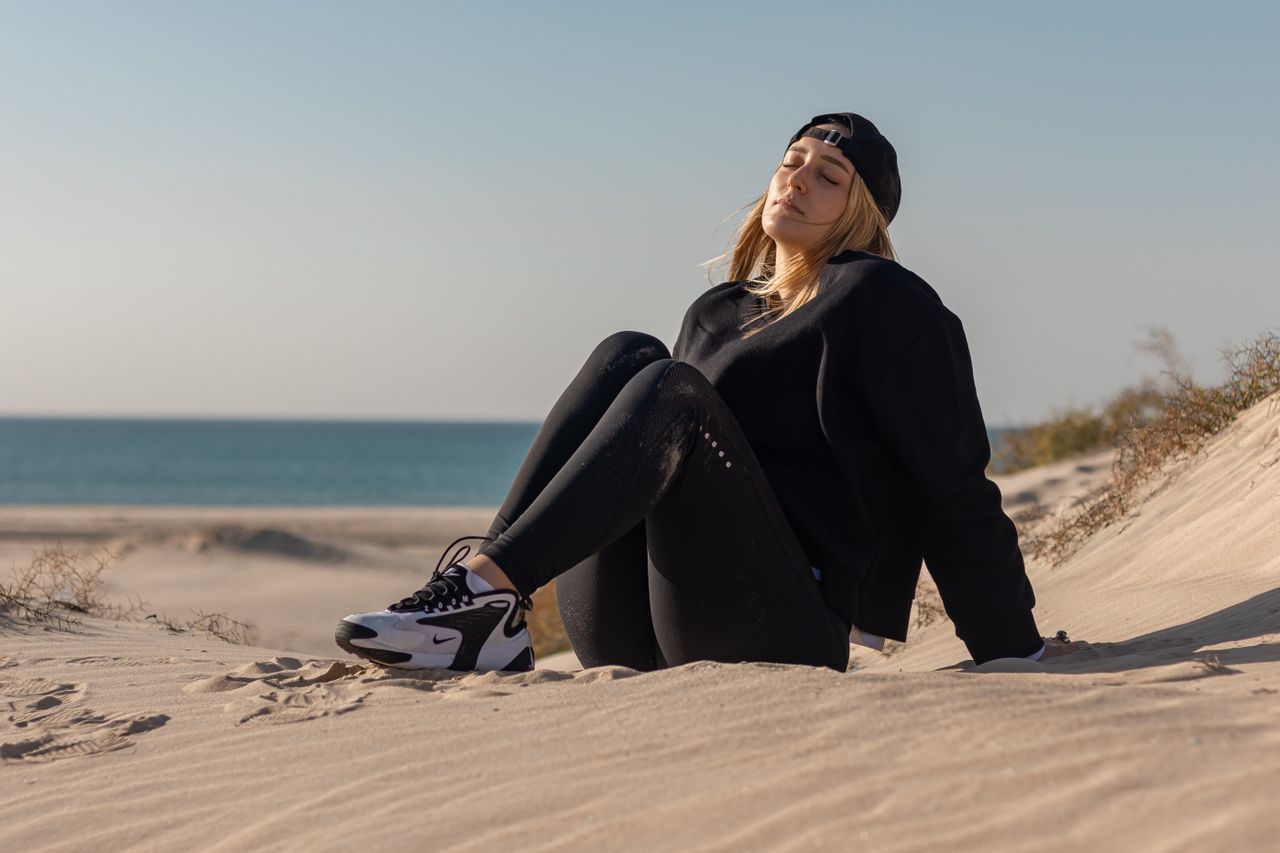 Full body of young blond female athlete wearing black activewear and cap sitting on sandy beach leaning on hands with closed eyes near sea under blue cloudless sky